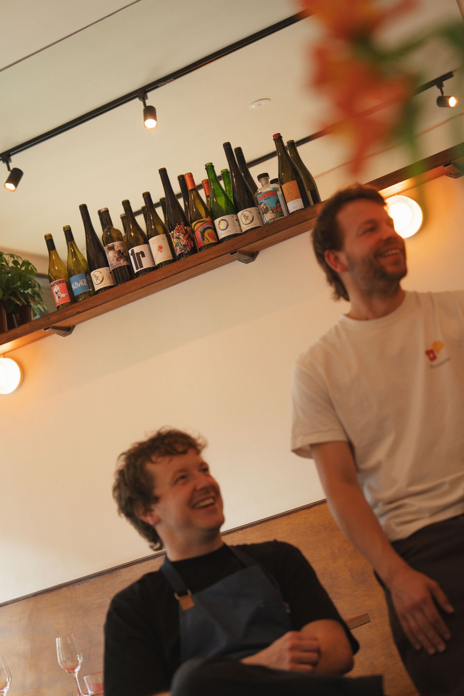 Two men laughing in a cozy restaurant with wine bottles displayed on a wooden shelf above them.