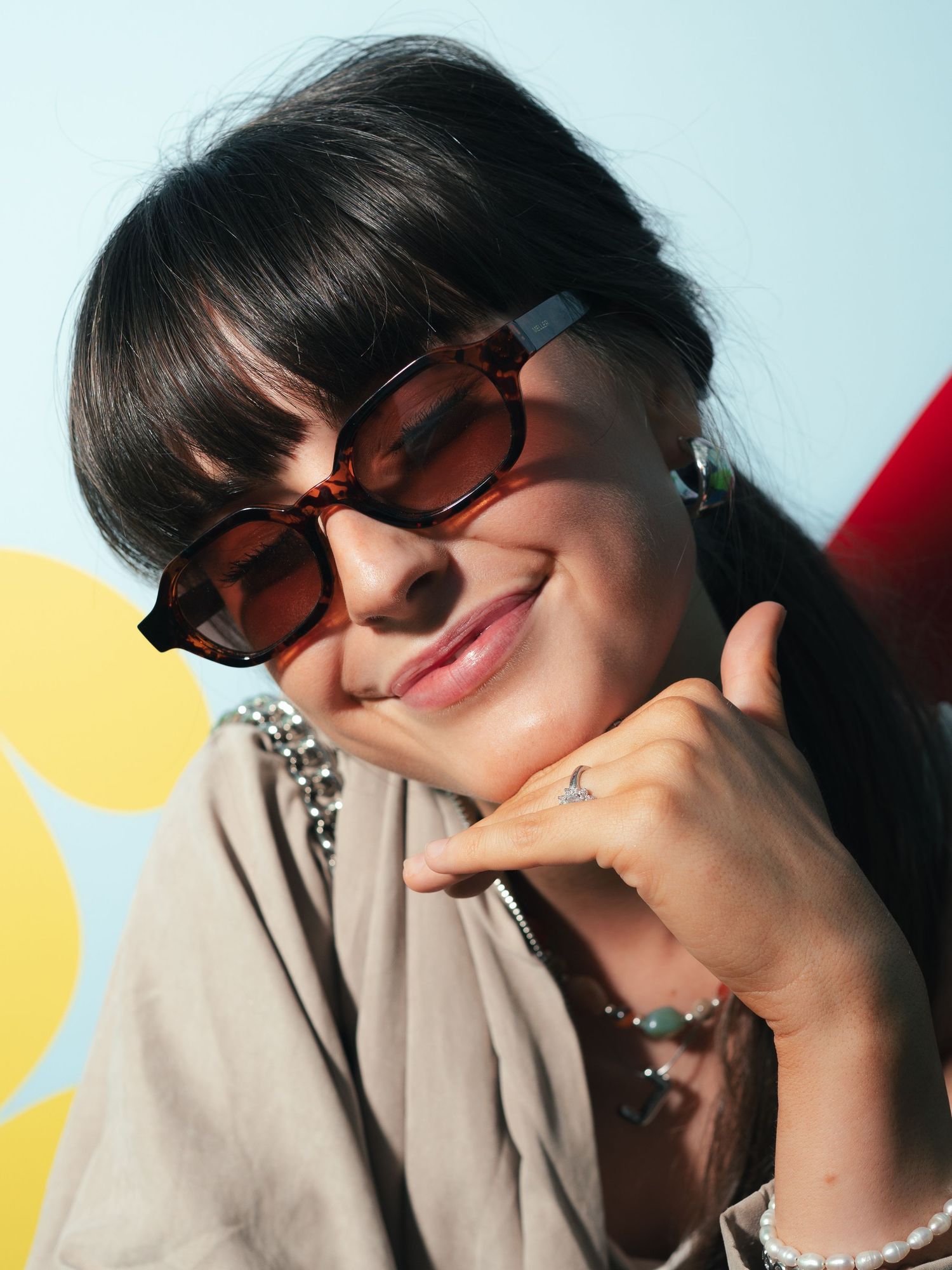 Woman with dark bangs wearing oversized brown tinted sunglasses and beige top smiling softly at an event.
