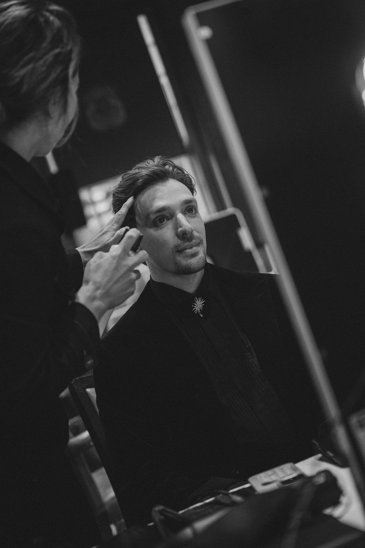 Black and white photo of a man getting makeup applied backstage, wearing a dark outfit with a brooch.