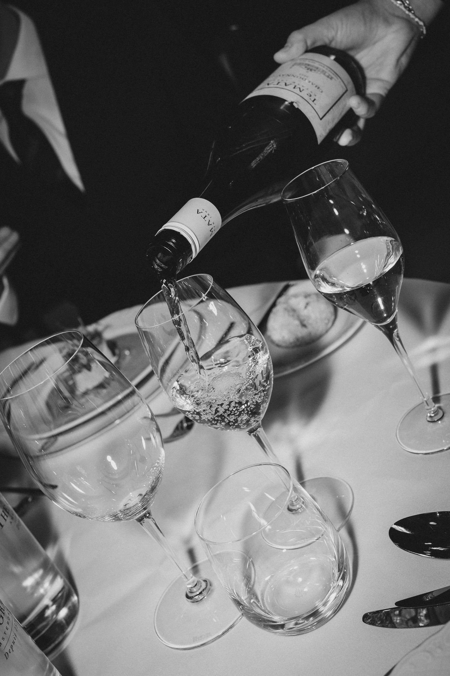 Black and white photo of wine being poured into crystal glasses on a white tablecloth at a formal dinner.