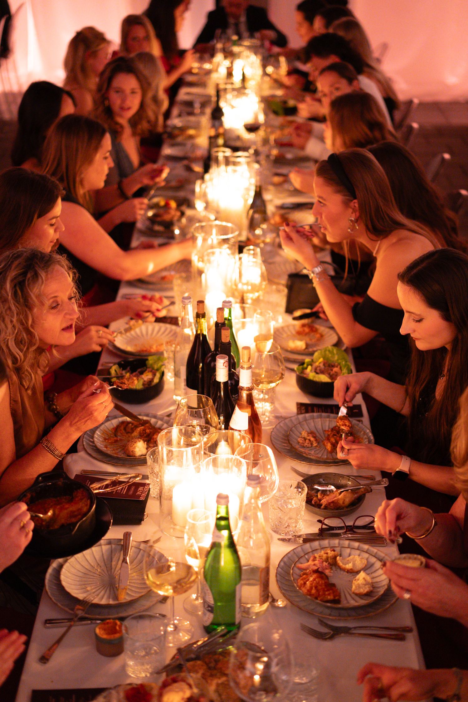 Women dining by candlelight at a long elegant dinner table with wine bottles, glasses, and plates of food.