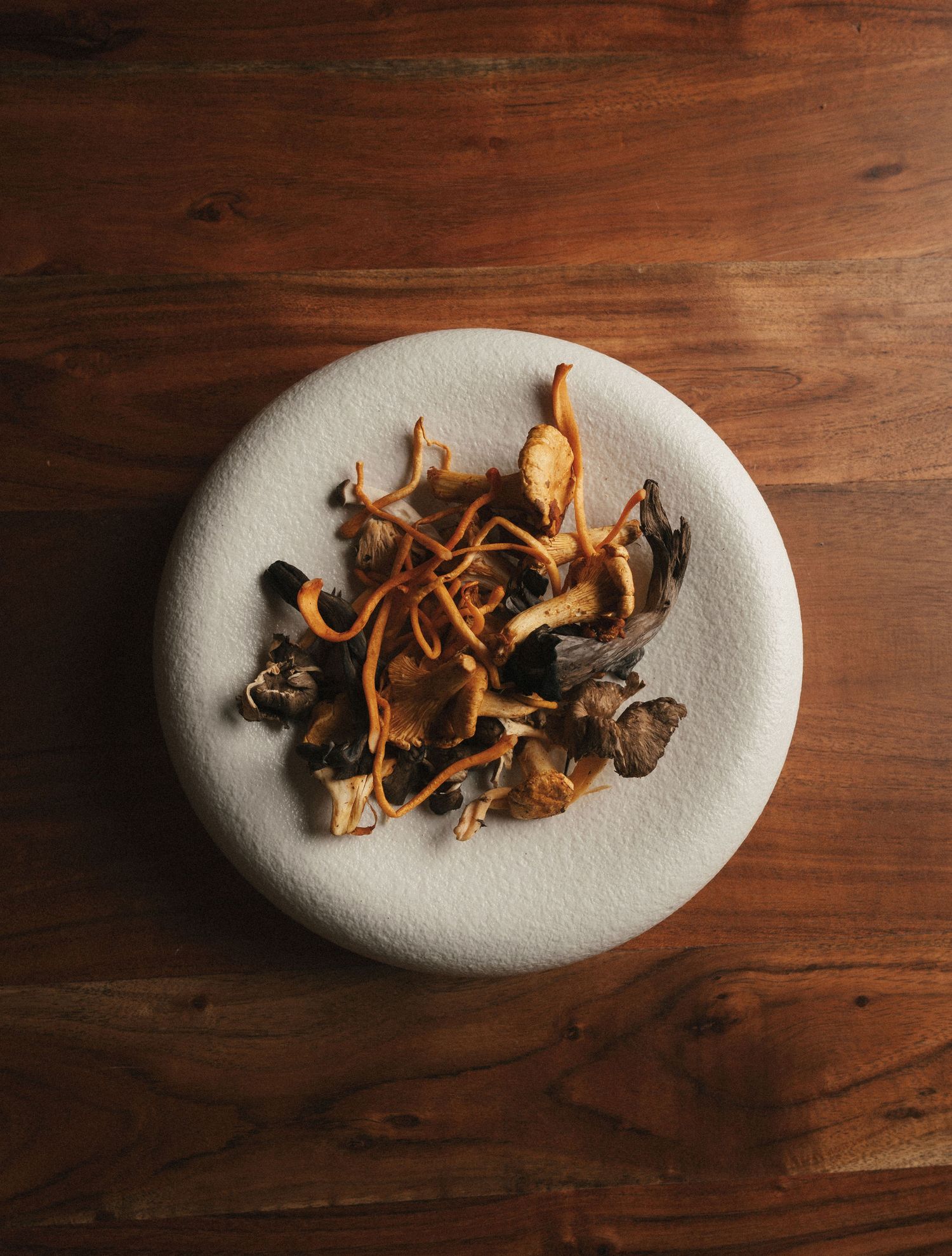 Dried mixed mushrooms with golden enoki on a white textured plate atop a rustic wooden table.