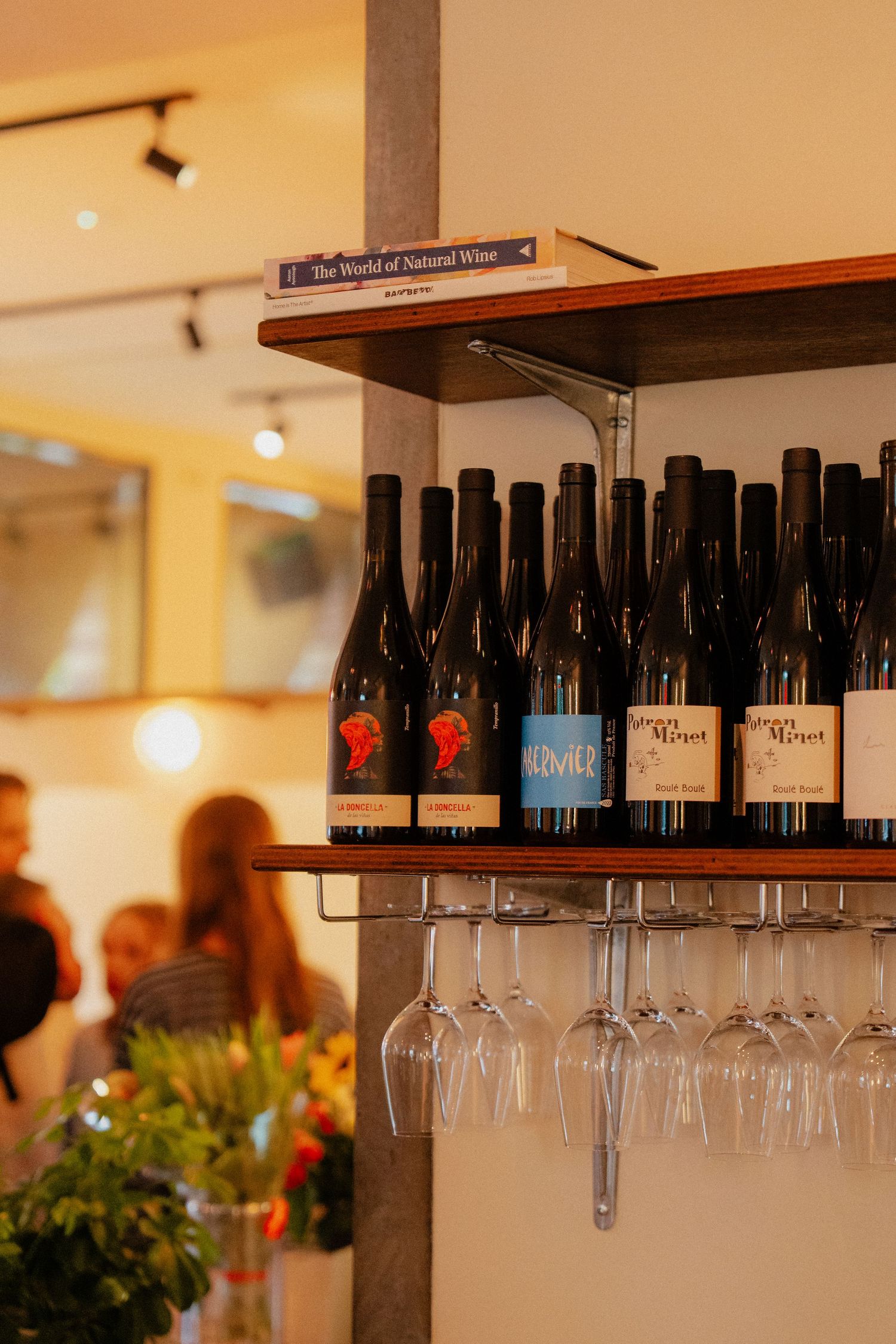 Wine bottles and glasses displayed on a wooden shelf in a cozy bar setting with guests in the background.