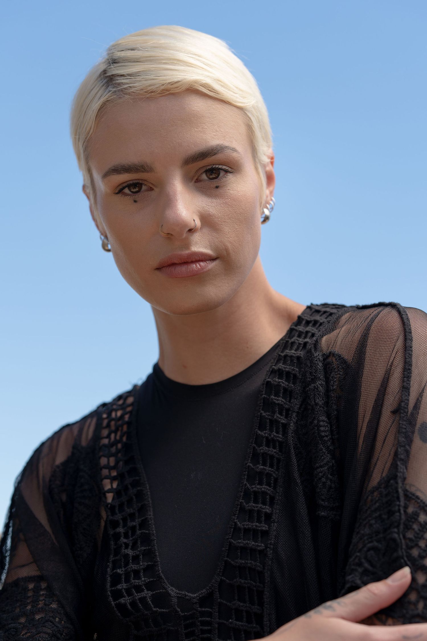 Person with short blonde hair wearing black lace top against blue sky background.