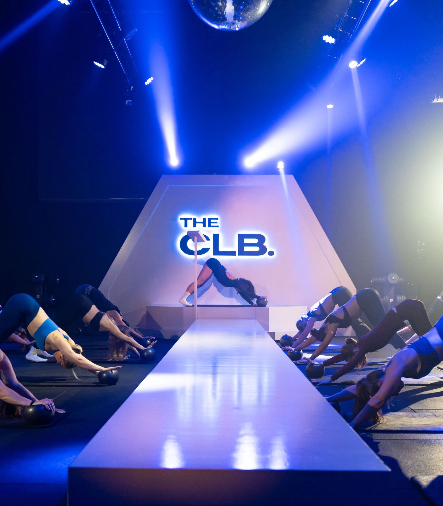 Group fitness class at The CLB studio with participants in downward dog pose under dramatic blue stage lighting.
