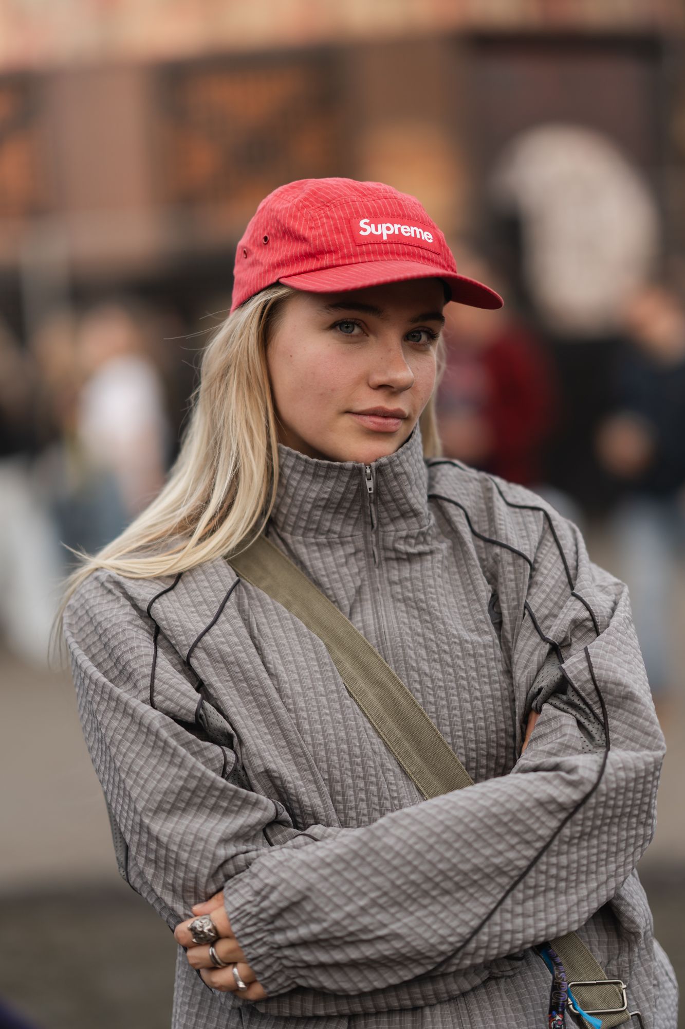 Young woman with blonde hair wearing a red Supreme cap and gray zip-up jacket, arms crossed outdoors.