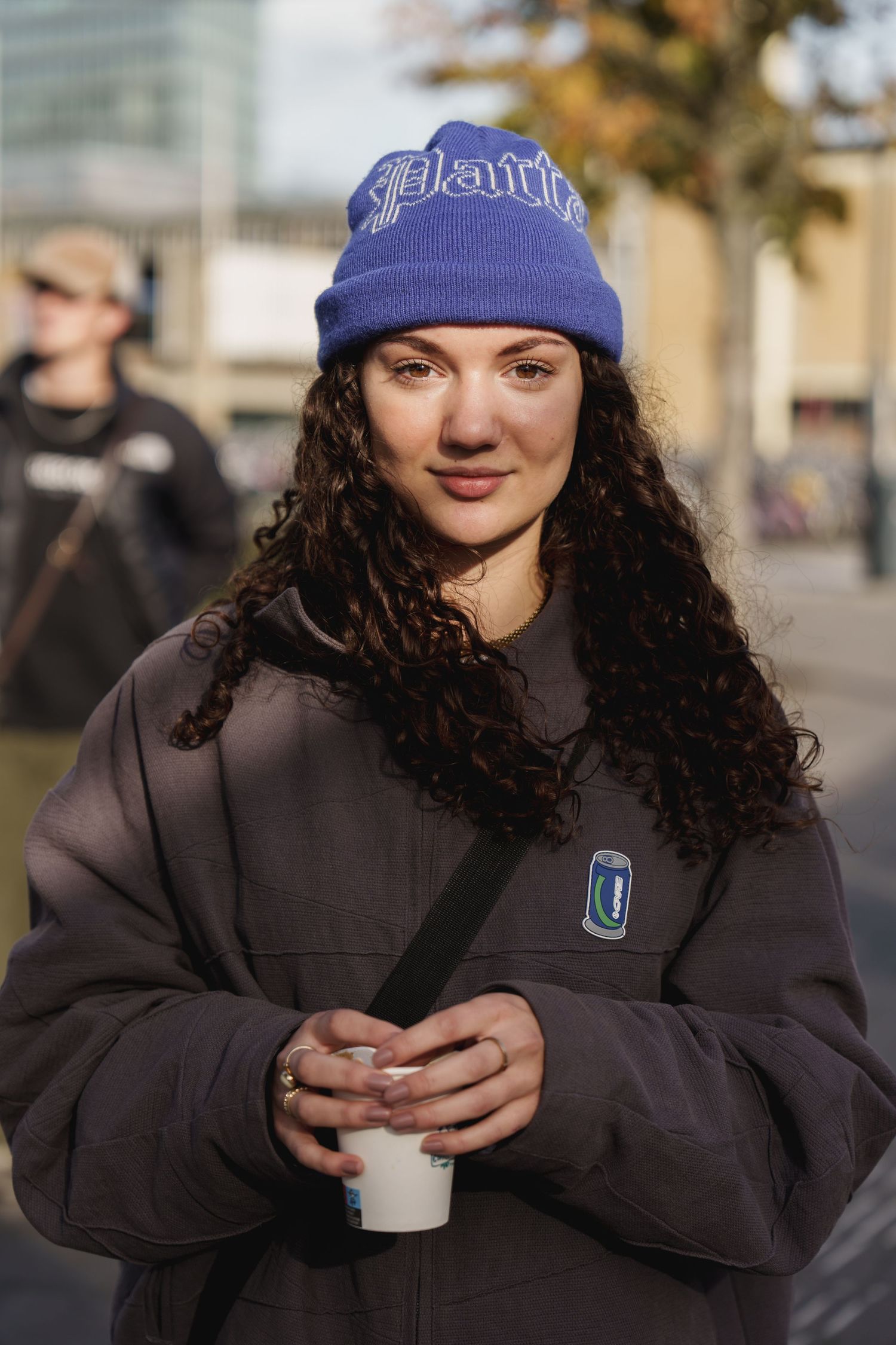 Young woman with curly hair wearing a blue beanie and gray hoodie, holding a coffee cup outdoors.