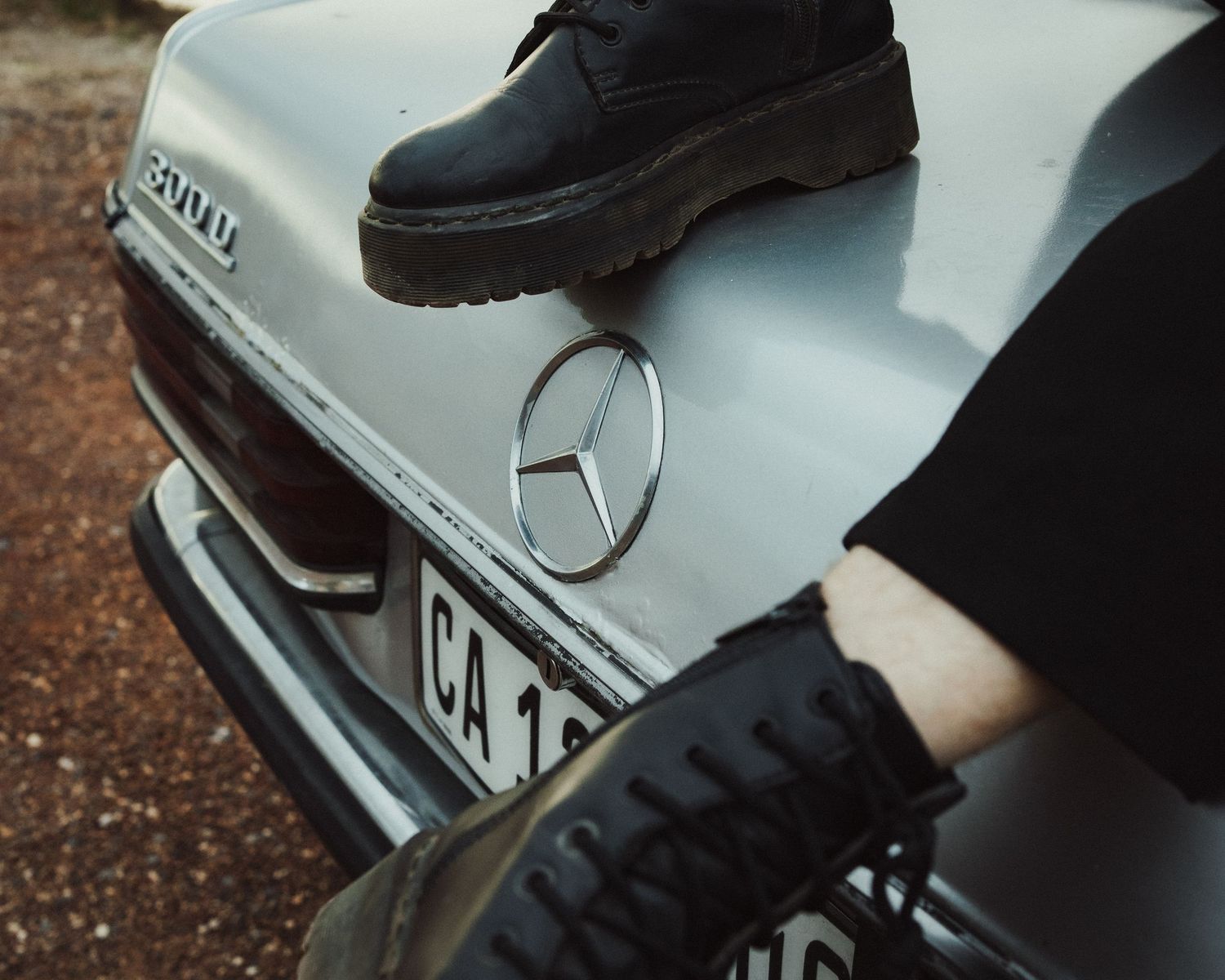 Person wearing black Dr. Martens boots resting on the hood of a vintage white Mercedes-Benz car with CA license plate.
