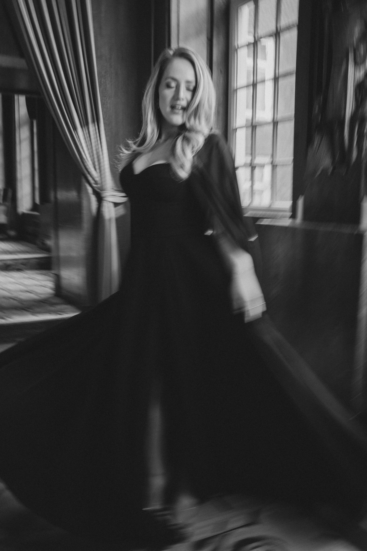 Black and white photo of a blonde woman in a flowing black gown twirling in a dimly lit vintage room.