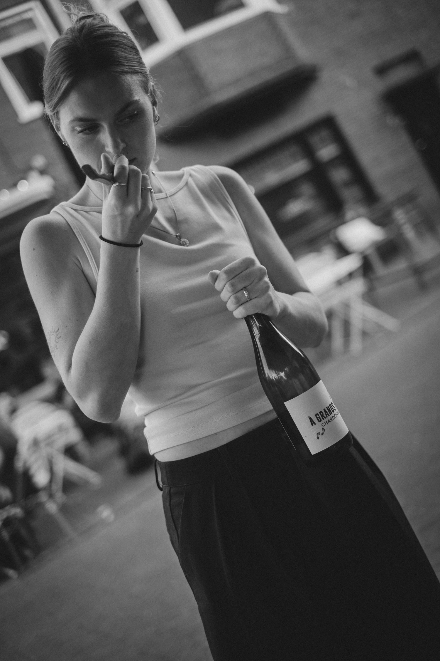 Woman in white sleeveless top holds wine bottle at outdoor restaurant in black and white photo.