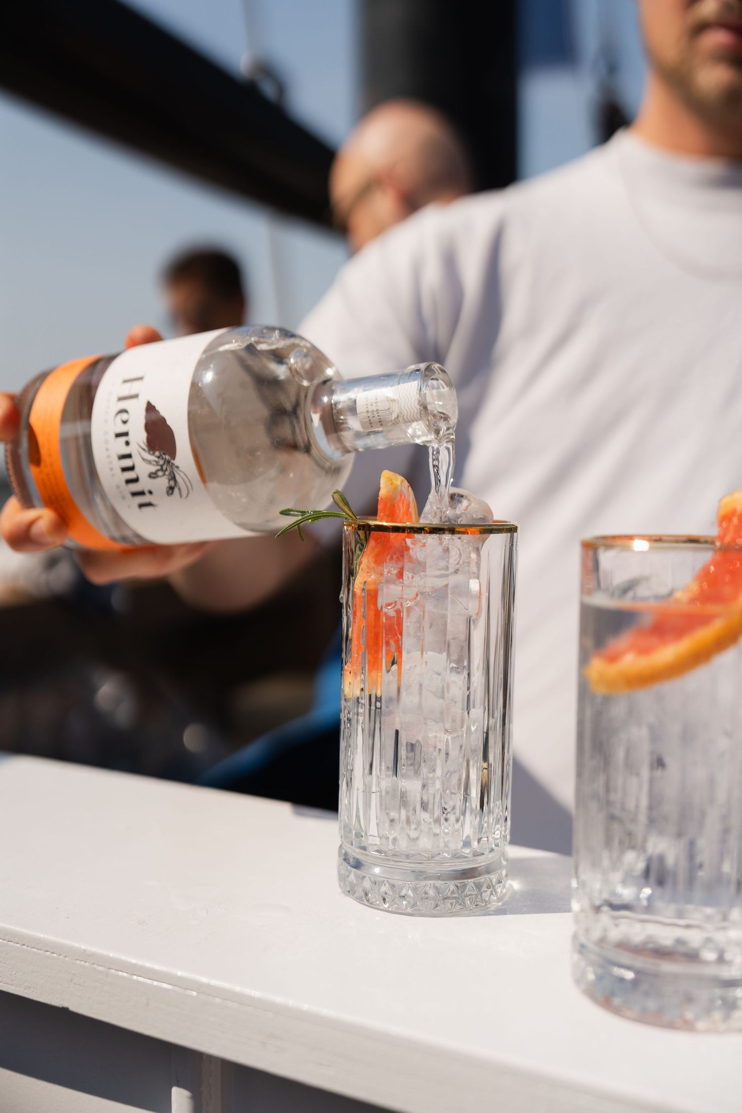 A bartender pours Hermit gin into a crystal glass garnished with grapefruit and rosemary on a sunny boat bar.