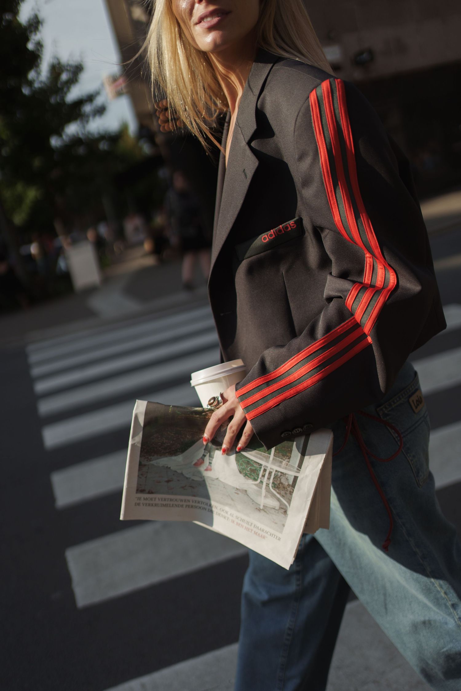 Blonde woman in striped blazer holding coffee cup and newspaper near crosswalk.