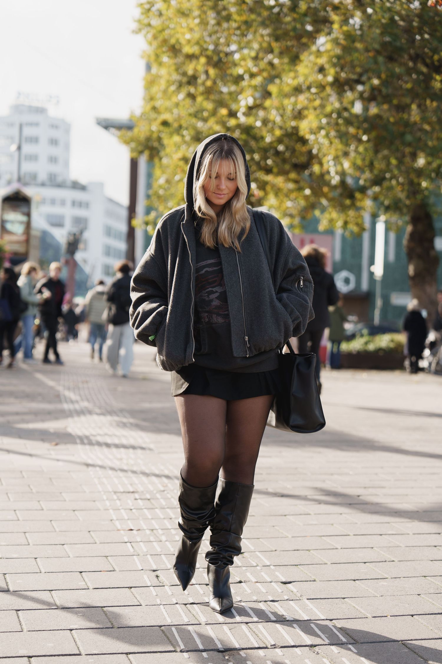 Woman in black oversized hoodie, mini shorts, tights and knee-high boots walking on a sunny city street.