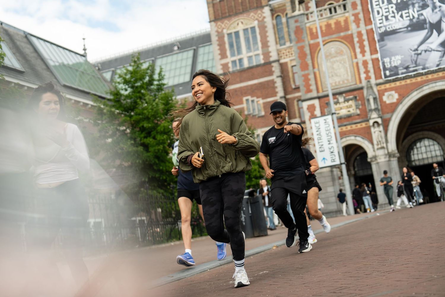 Woman jogging in green jacket near historic brick building in Amsterdam, with people walking in background.