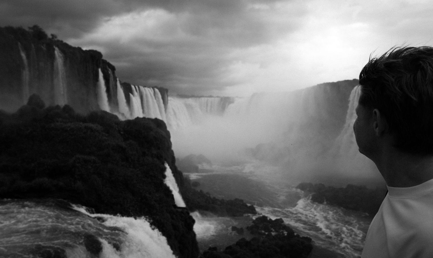 Black and white photo of a person gazing at the majestic Iguazu Falls surrounded by mist and dramatic skies.