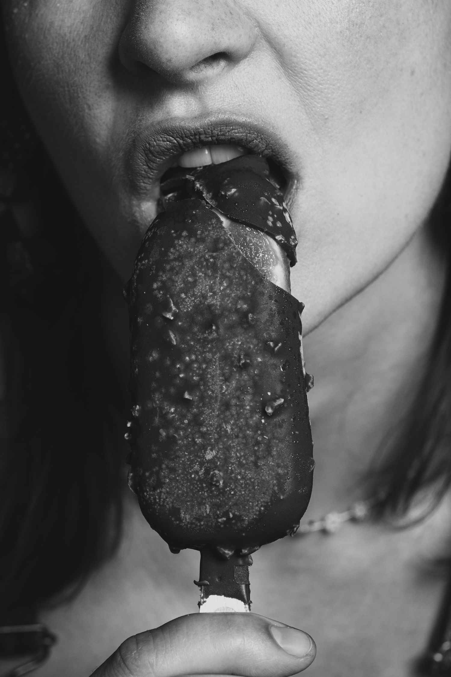 Black and white close-up of a woman biting into a chocolate-covered ice cream bar on a stick.