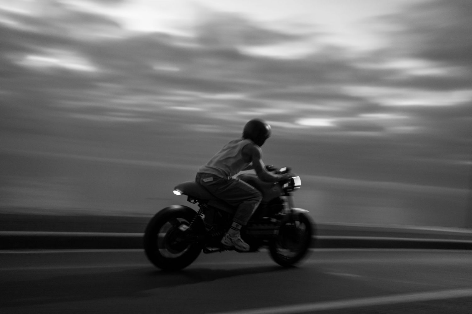 Black and white panning shot of a motorcyclist riding at high speed on an open road under dramatic cloudy skies.