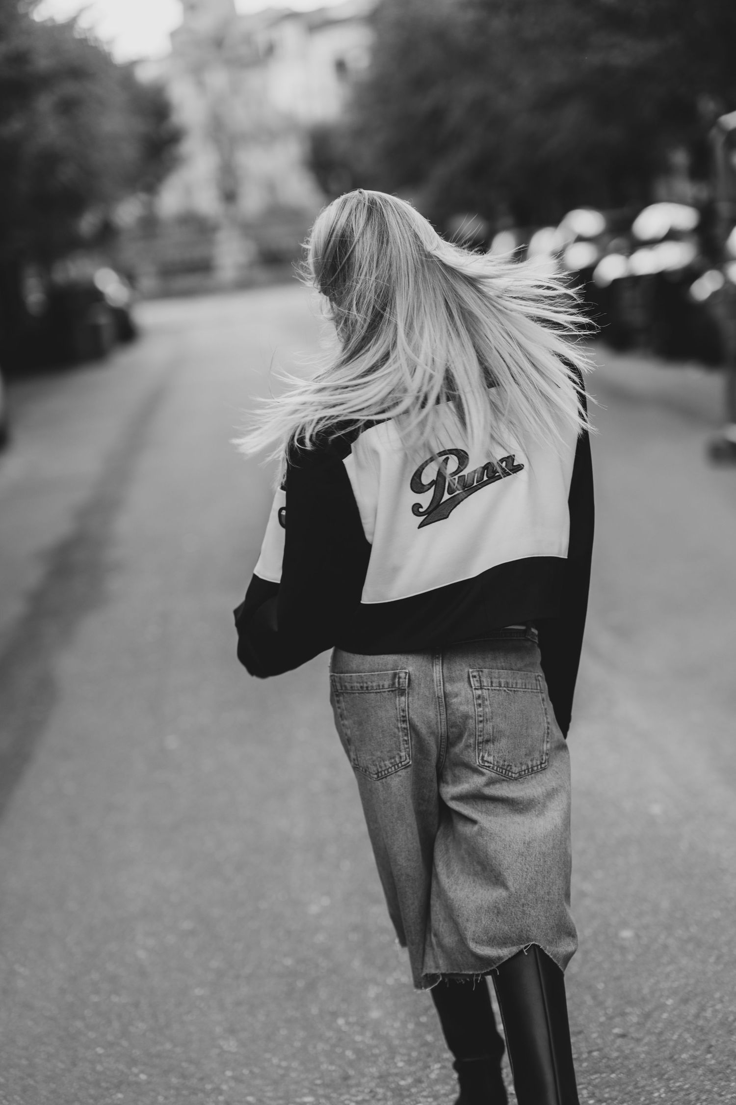 Black and white photo of a blonde woman walking away in a Puma varsity jacket and jeans on a city street.