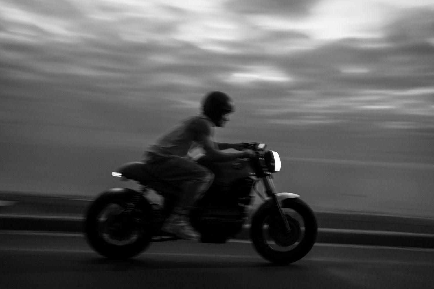 Black and white panning shot of a motorcyclist speeding on a road with a dramatic cloudy sky backdrop.