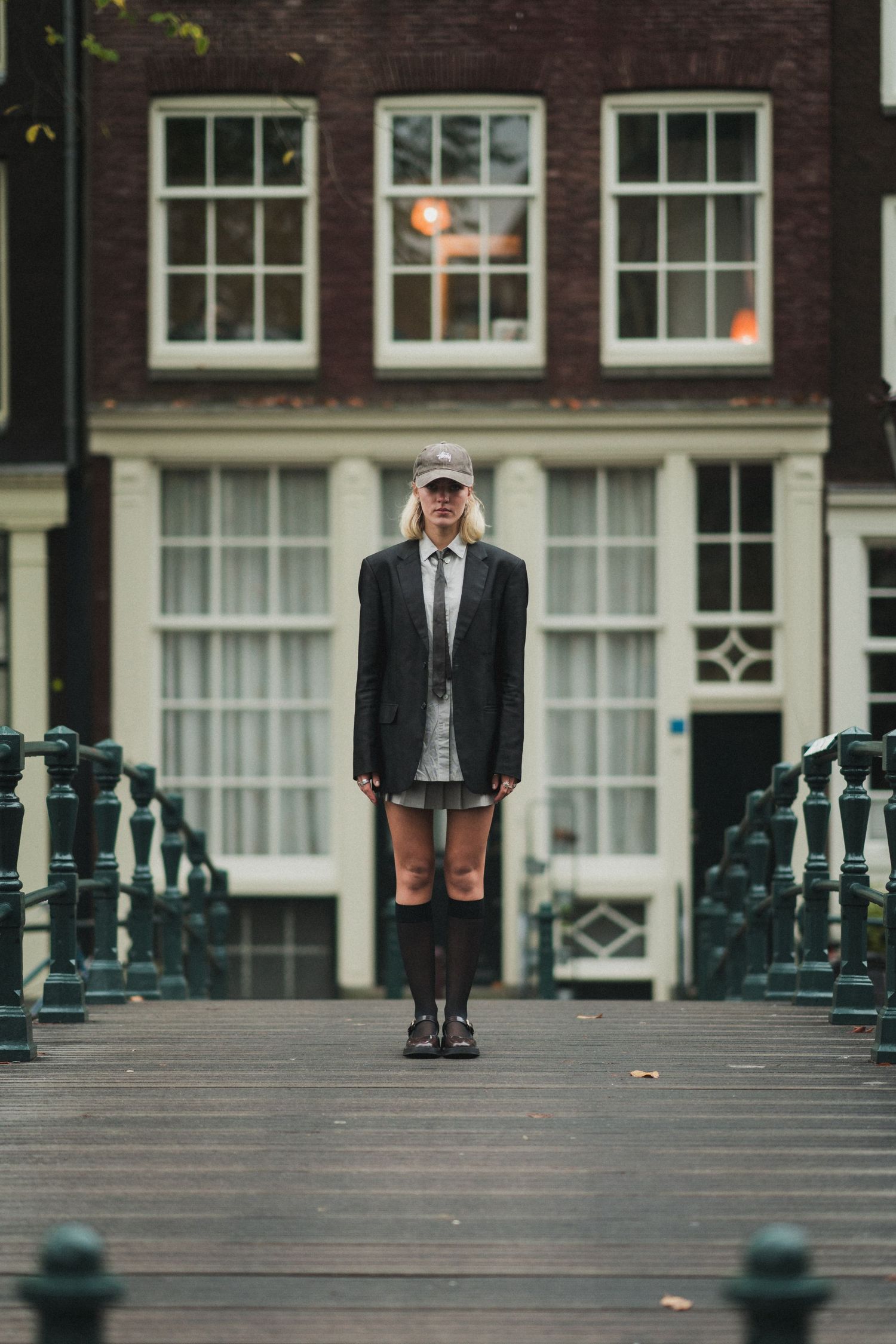 A blonde woman in a blazer, tie, cap, and knee socks stands on a wooden bridge in Amsterdam.