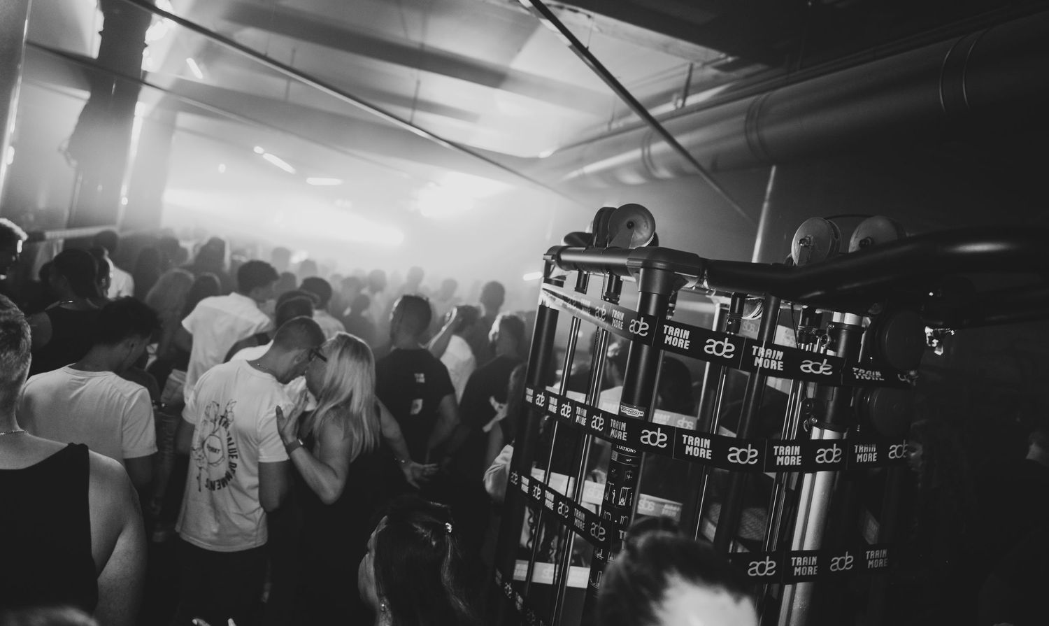 Black and white photo of a crowded nightclub with fog, dancers, and DJ equipment visible.