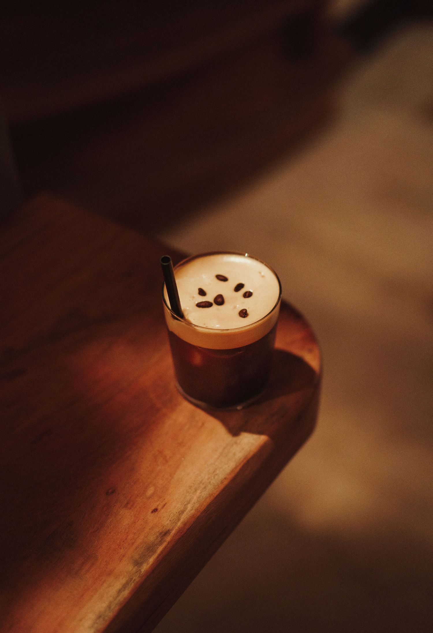A frothy cappuccino with coffee beans on top sits in a glass cup on a warm wooden table in soft light.