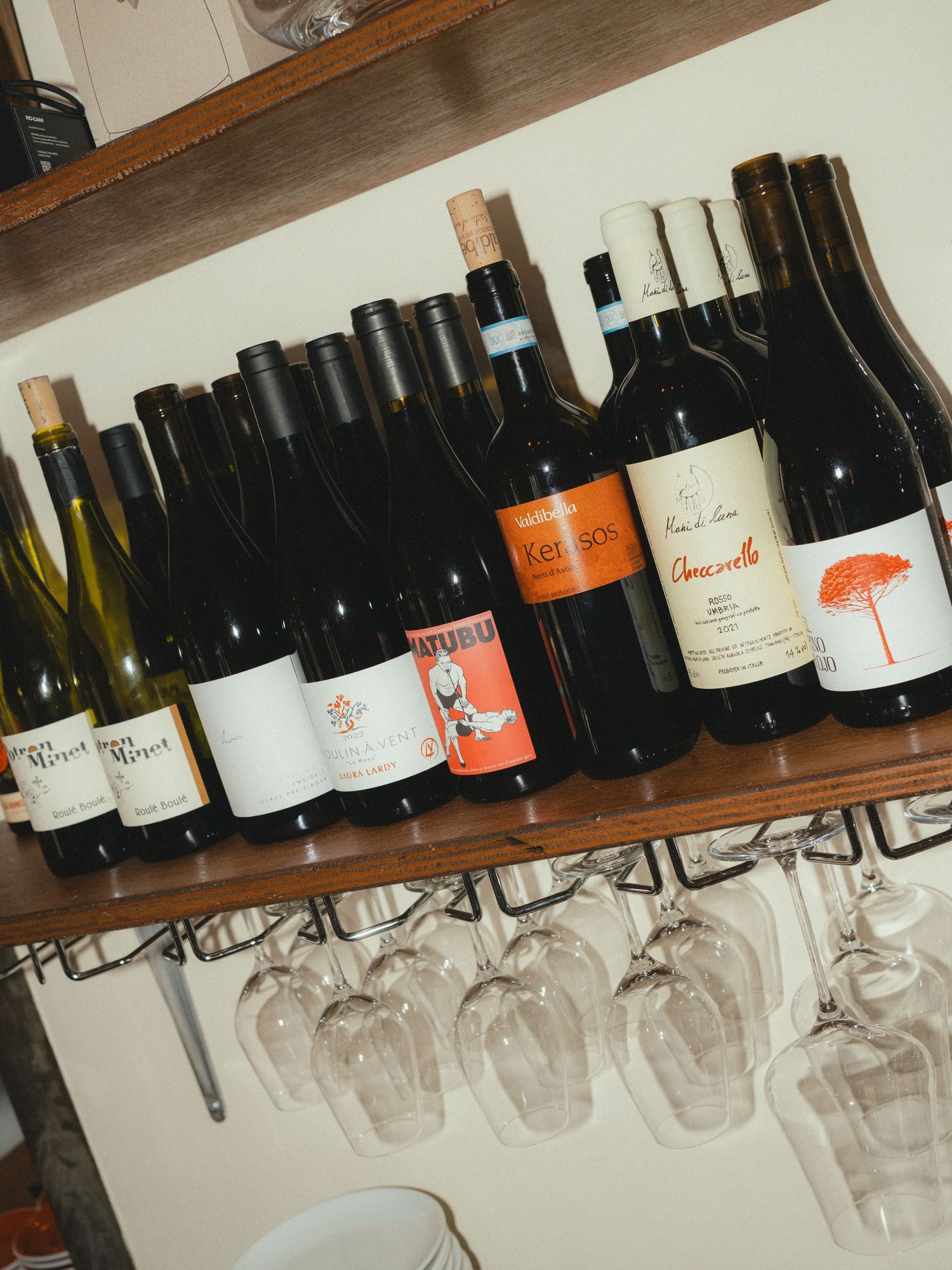 A wooden shelf displaying an assortment of wine bottles with hanging wine glasses underneath.