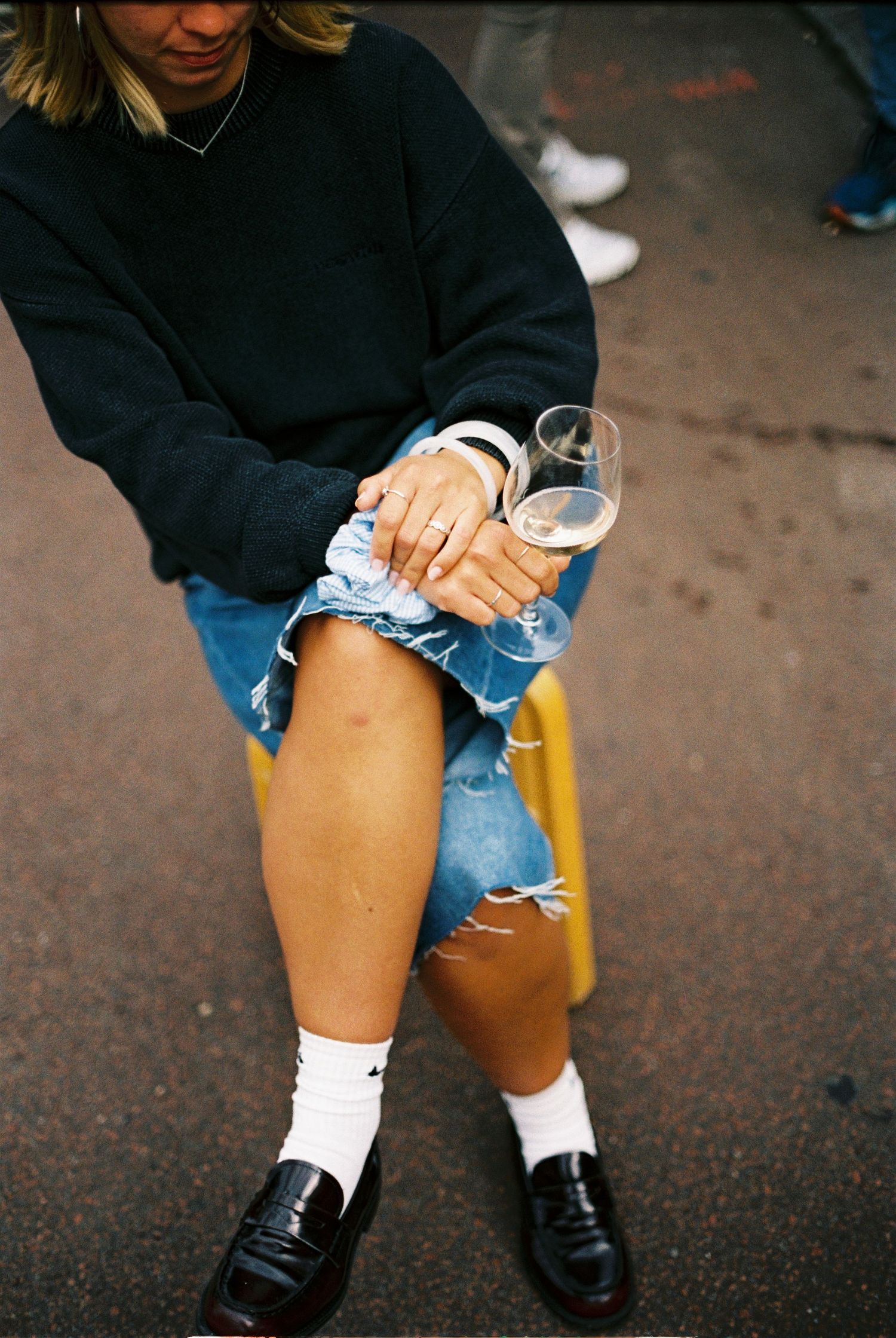 Woman in black sweatshirt and denim mini skirt holding a wine glass while seated outdoors.
