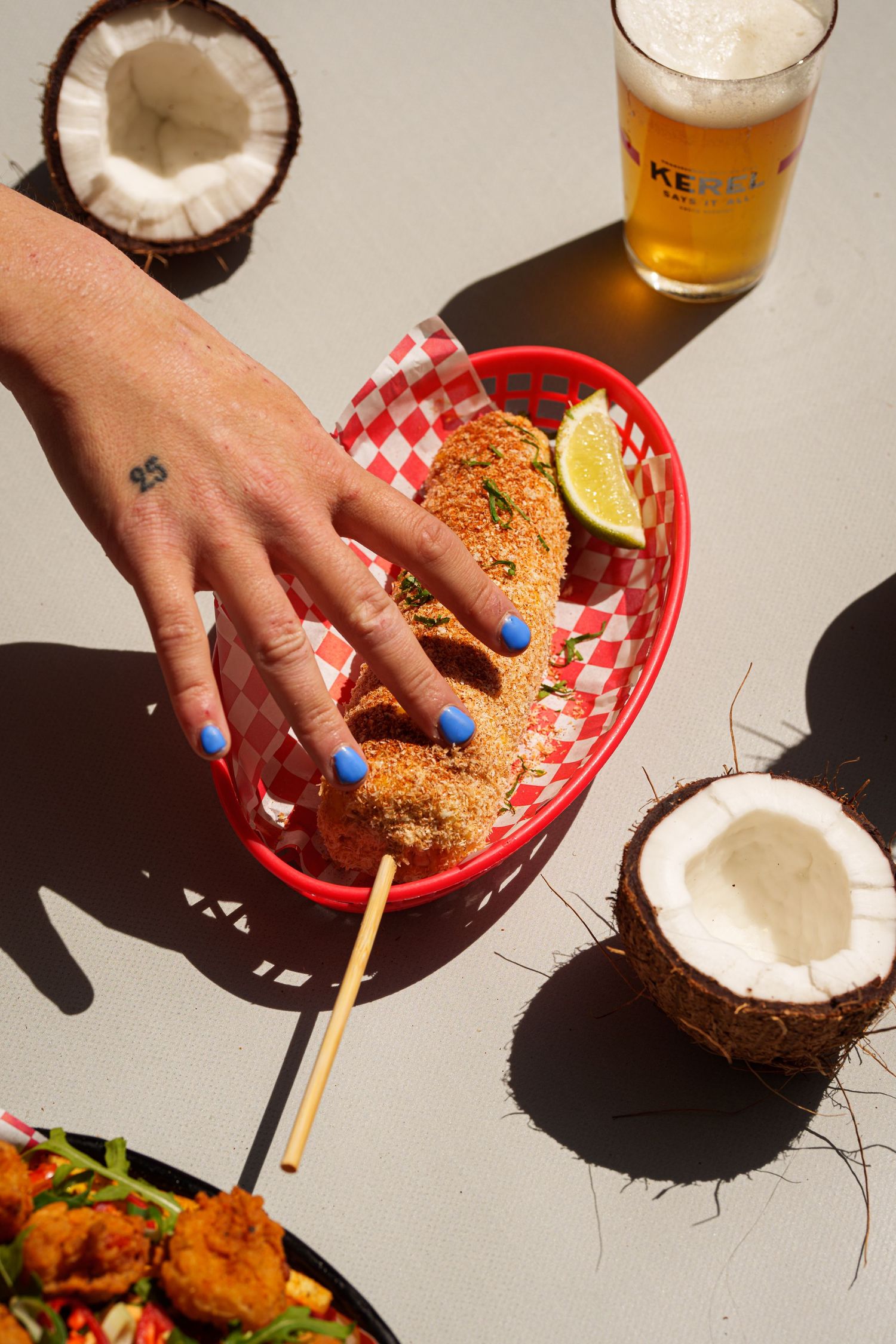 Hand with blue nails reaching for breaded food in red basket, with coconuts and beer on table.