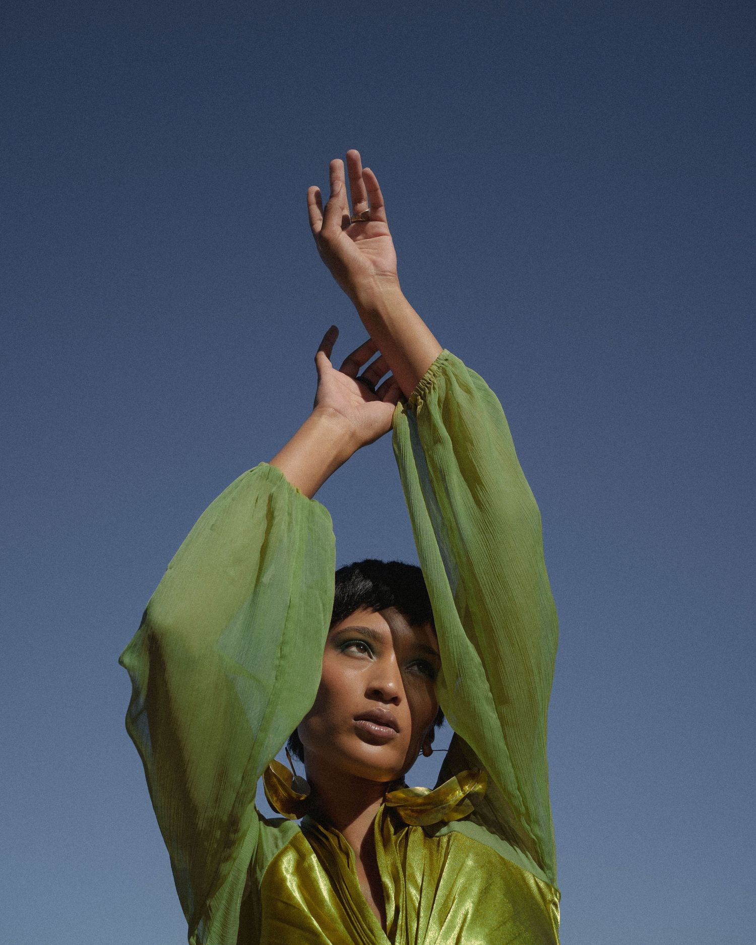 Woman in green satin kaftan raises arms against blue sky in artistic fashion pose.