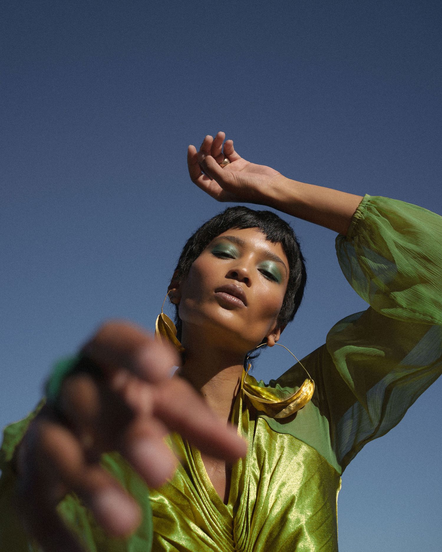 Woman in green metallic outfit with gold necklace and green eyeshadow poses against blue sky.