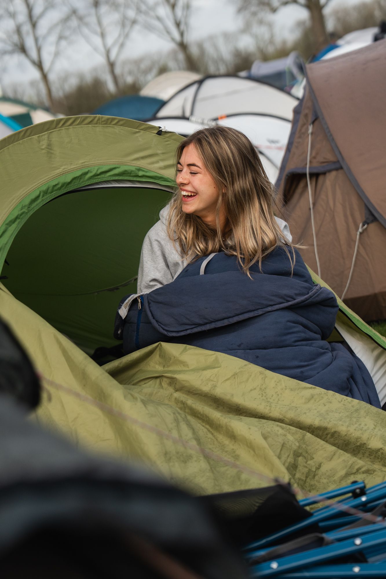 Young woman laughing joyfully while sitting in a green tent at a campsite, wrapped in a sleeping bag.