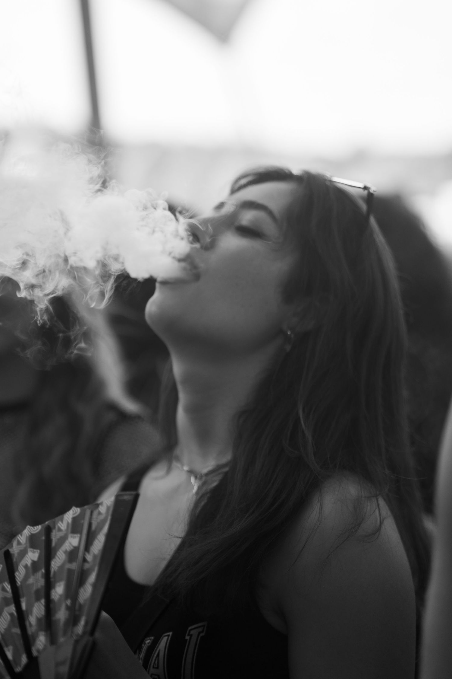 Black and white photo of a young woman exhaling a large cloud of smoke, holding a decorative fan outdoors.