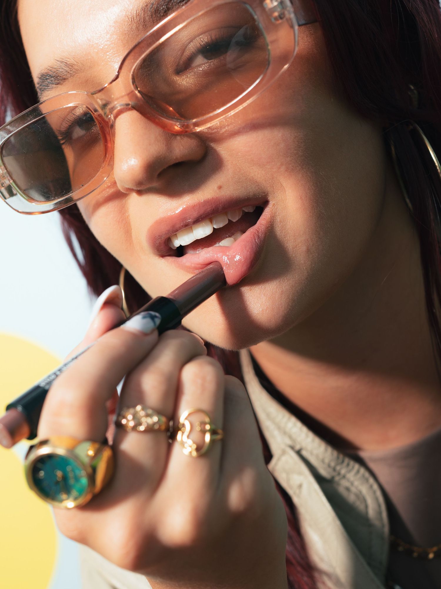 Woman applying nude lipstick wearing pink sunglasses and gold rings.