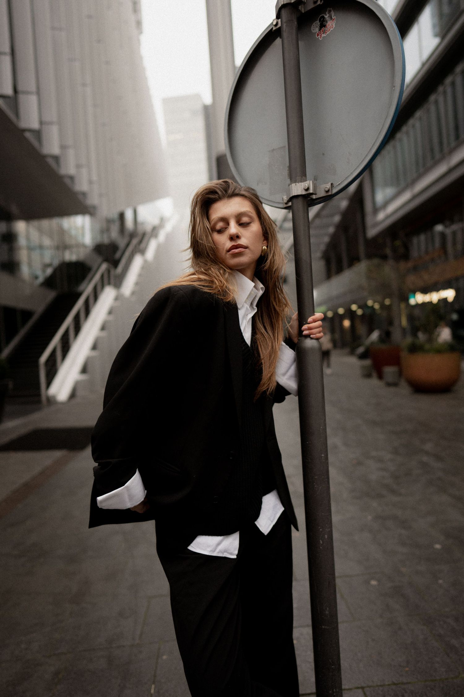 Young woman in black suit leaning against a street sign pole in an urban city setting.