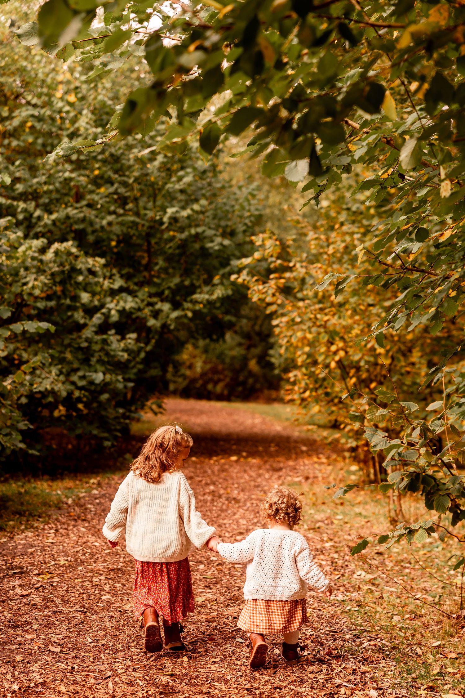 Two small children walking together on autumn path covered in fallen leaves.