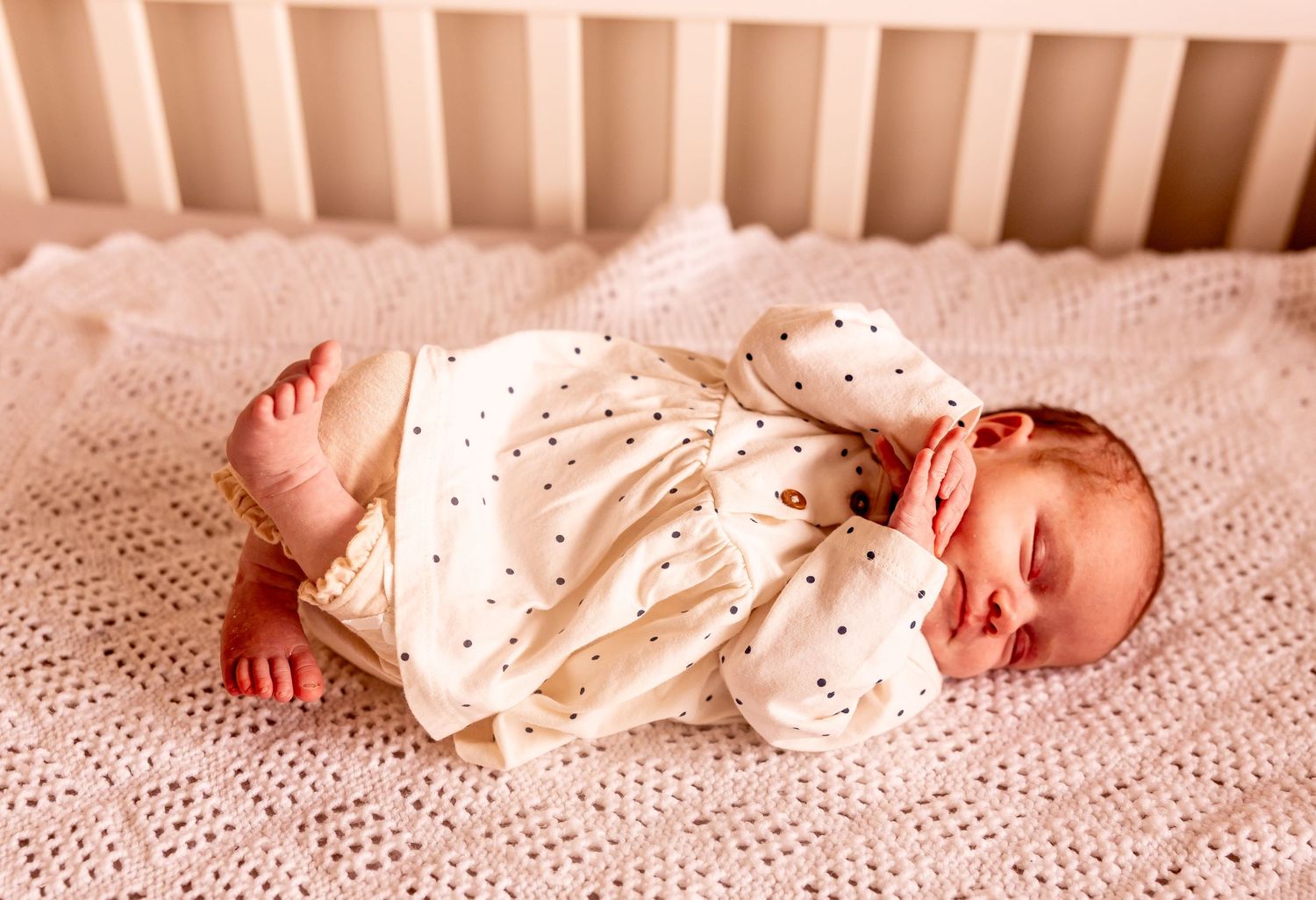 A smiling baby wearing a polka dot outfit lying on a pink blanket in a crib.