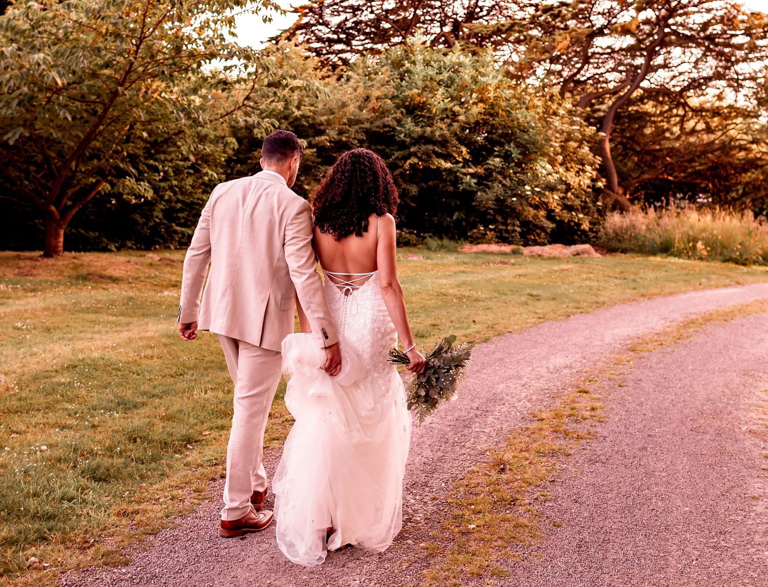 A couple walks hand in hand down a countryside path at sunset in wedding attire.
