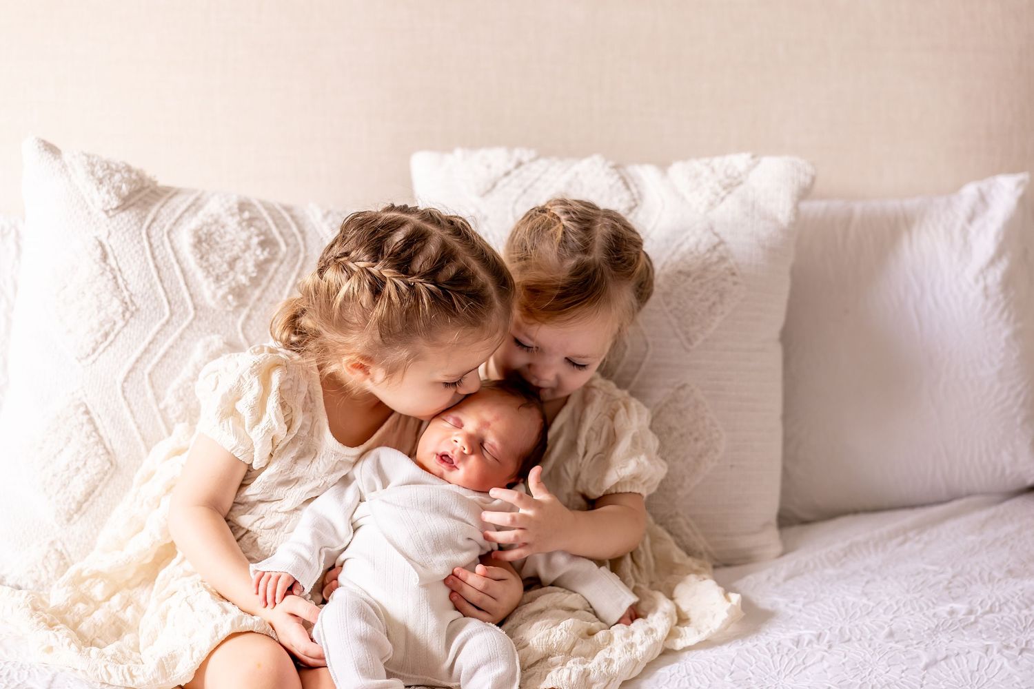 Two young children cuddling with newborn sibling on white bedding.