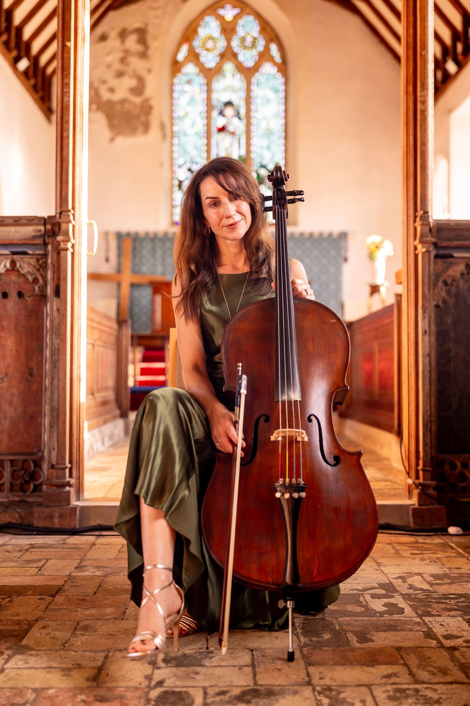 Musician poses with cello in historic church setting with stained glass windows.