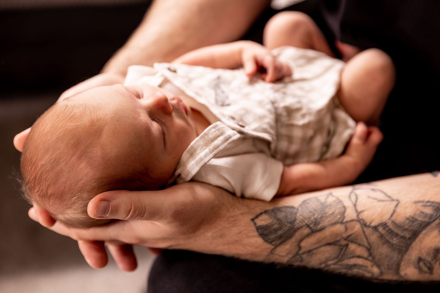 Tattooed arms carefully cradling sleeping newborn in white clothing.