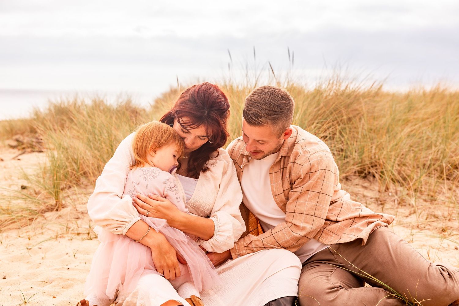 Family sitting together on sandy beach dunes with newborn.