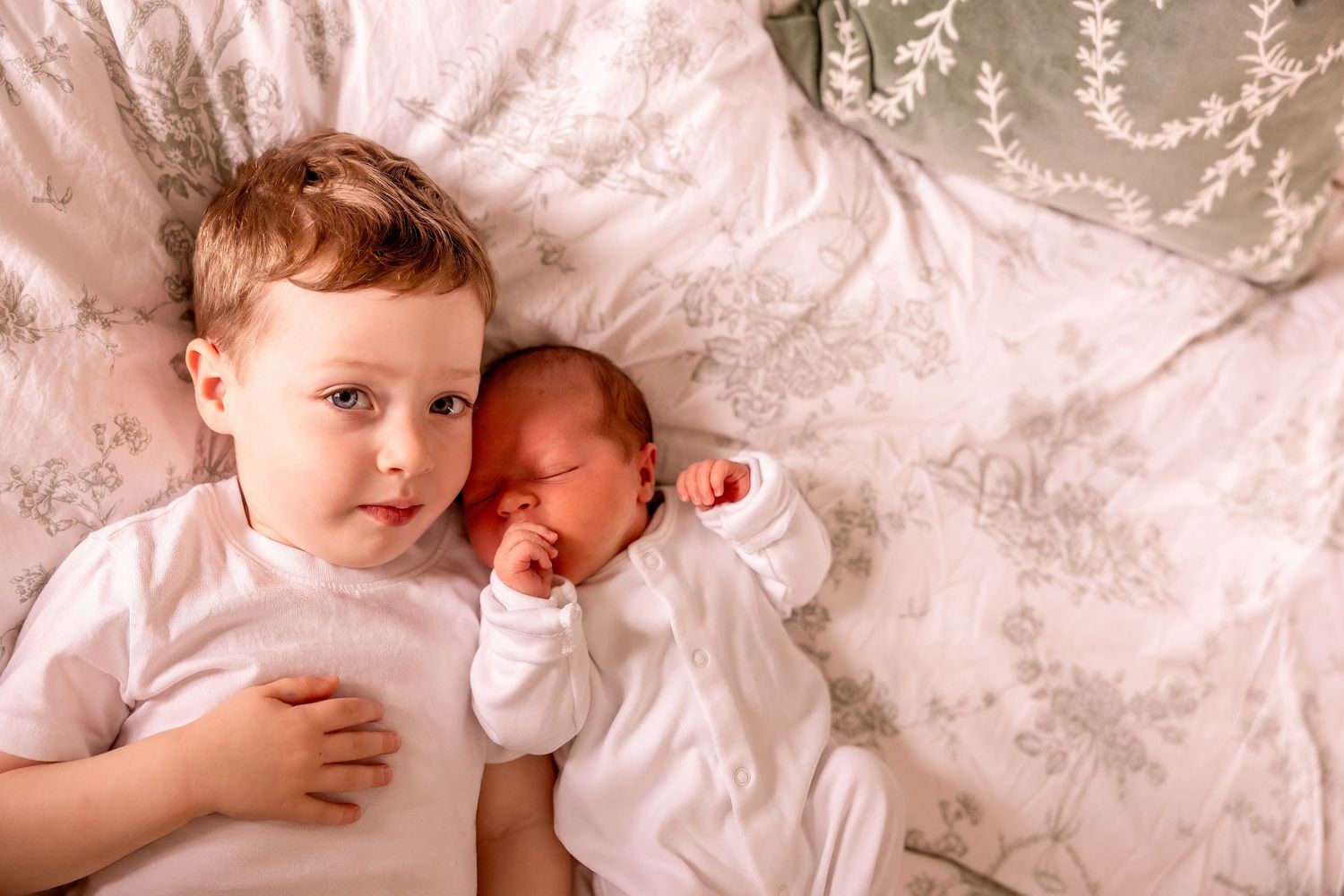 Two siblings lying on floral bedding with newborn baby in white outfit.