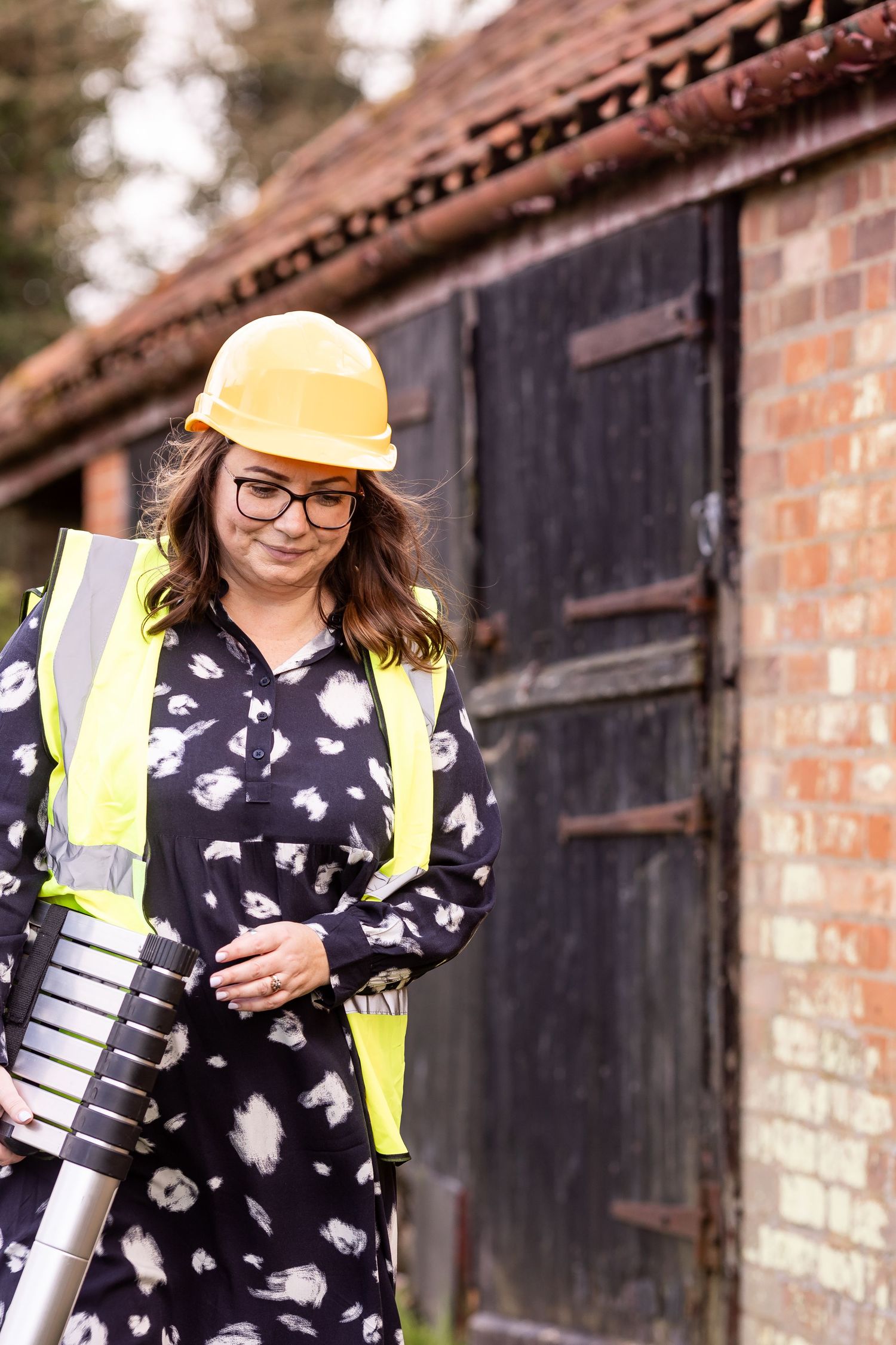 A person in a floral print dress and safety vest stands against a brick wall background.
