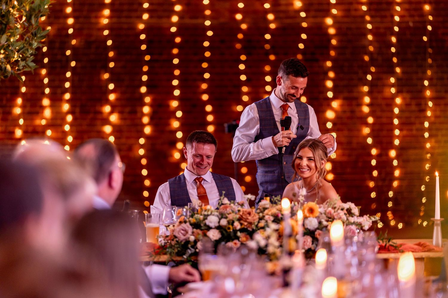Wedding reception venue decorated with hanging string lights and floral centerpieces during evening toast.