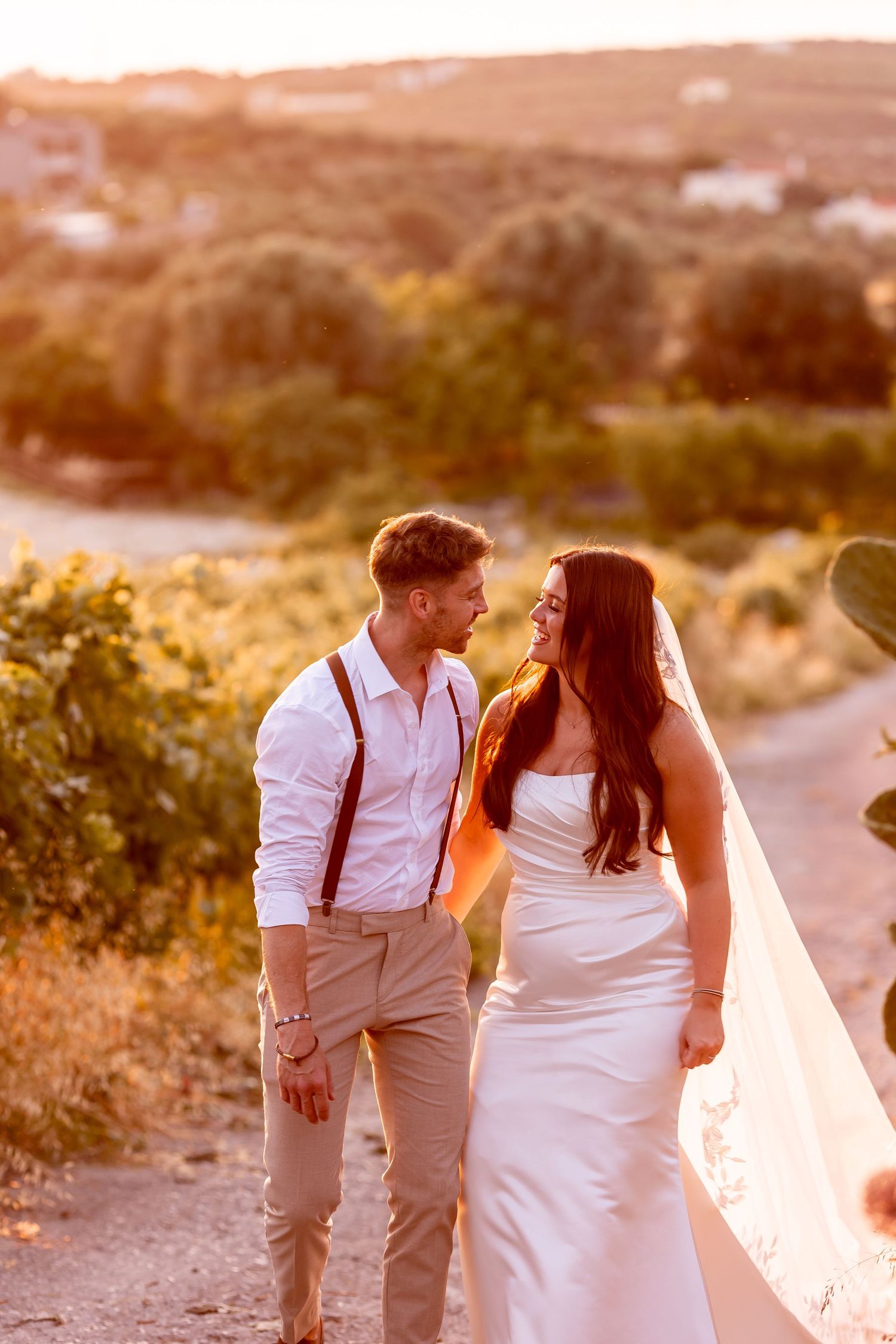 Newlyweds in white attire walking together during golden hour with long flowing veil.