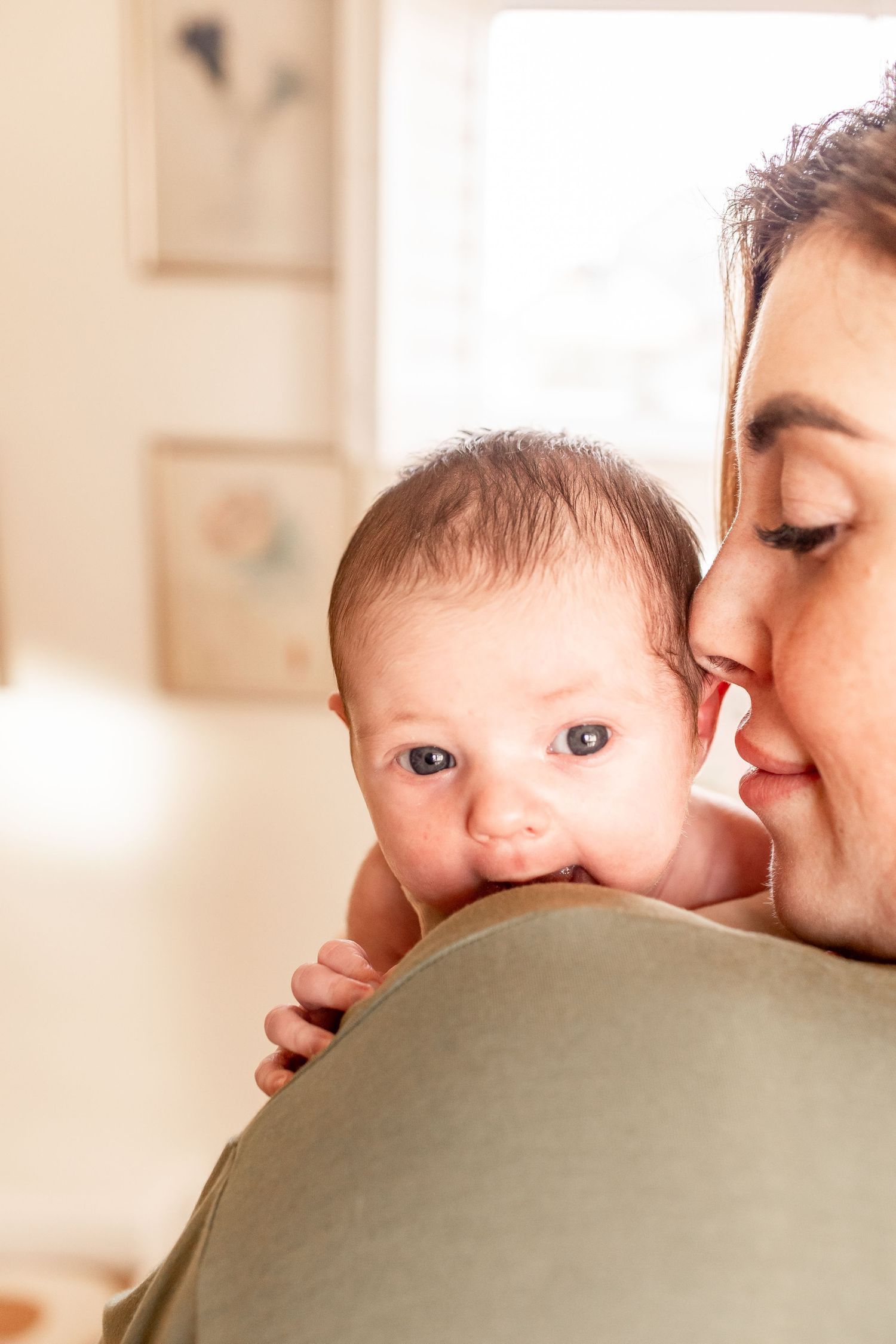 Baby peering over caregiver's shoulder in warm sunlit room.
