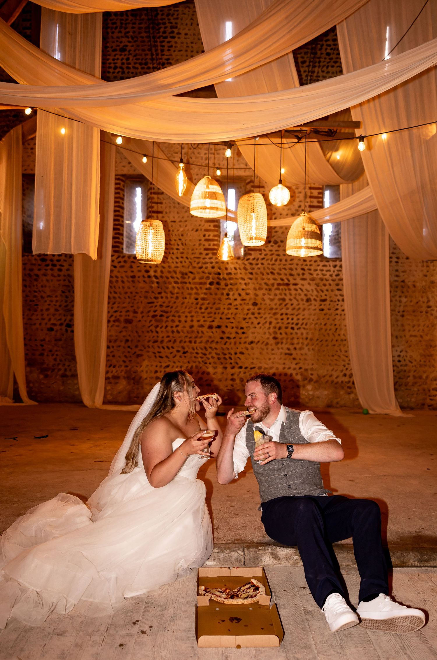 Wedding couple shares playful moments on the floor of a rustic venue decorated with flowing white fabric and string lights.