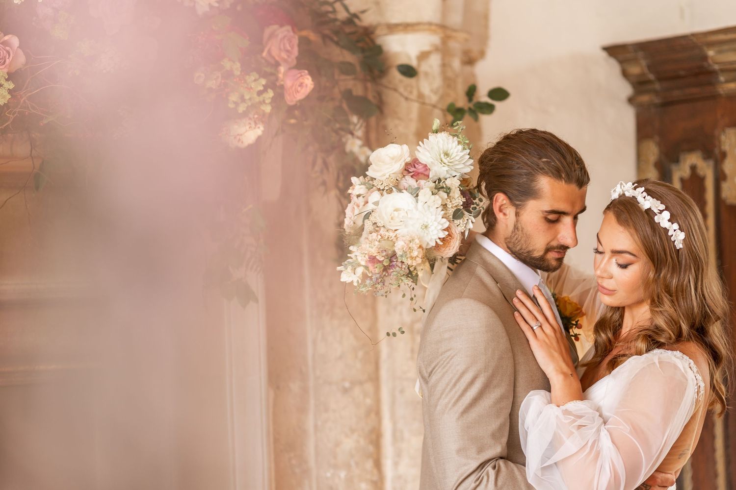 Romantic wedding couple embraces in soft pink light surrounded by delicate floral decorations.