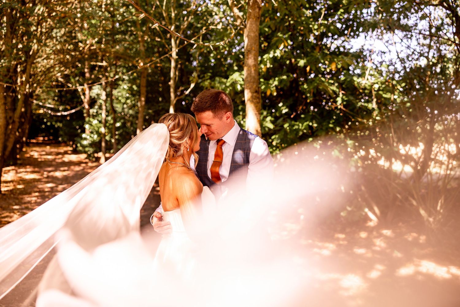 Wedding couple embraces outdoors as sunlight filters through bride's flowing veil in a wooded setting.